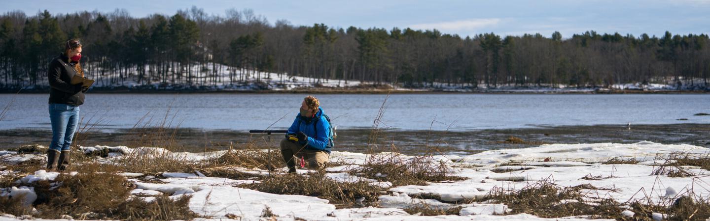 Two coastal UNH researchers work at the edge of an estuary in winter, with snow in the foreground